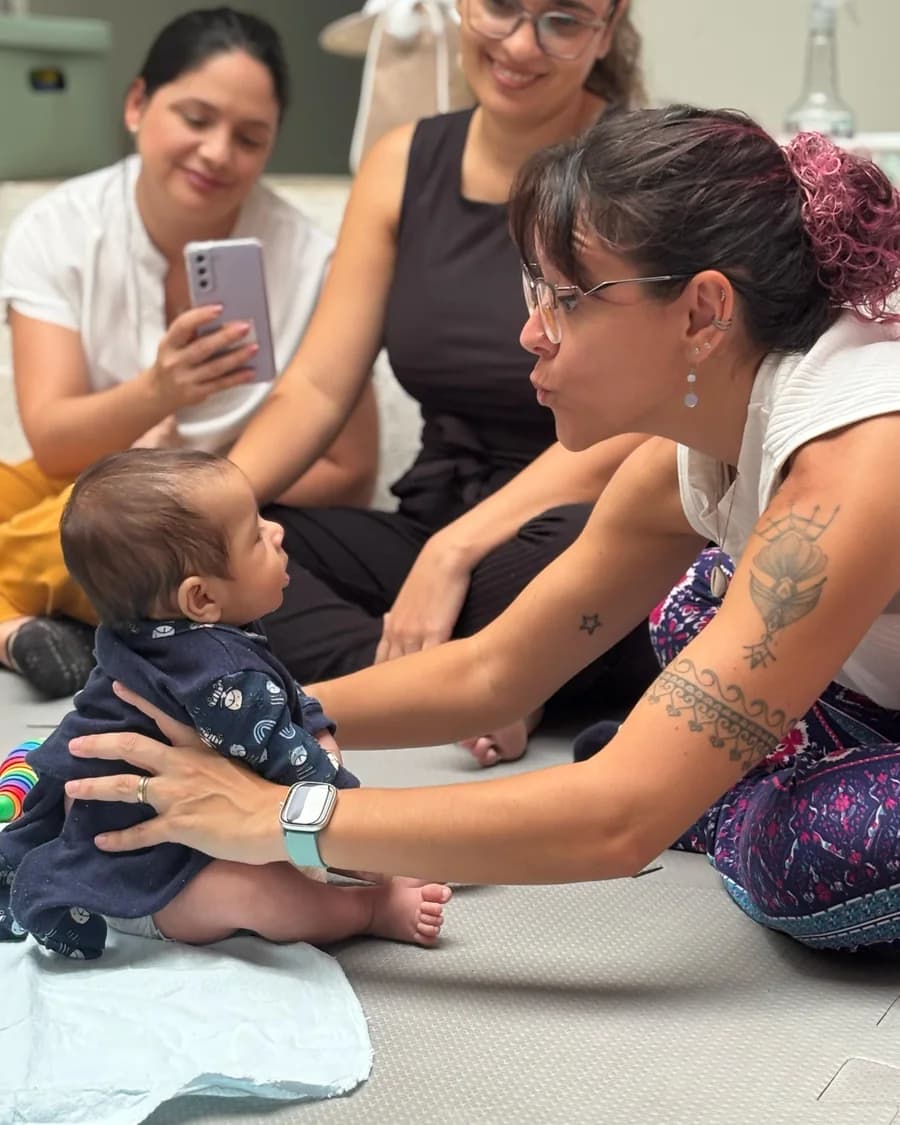 Profª Drª Dafne Herrero em contato visual com um bebê, apoiando-o sentado no colchonete, enquanto a mãe registra a cena