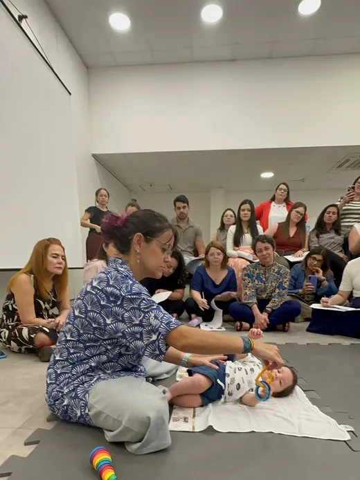 Dafne atendendo um bebê no chão diante de uma turma sentada em roda