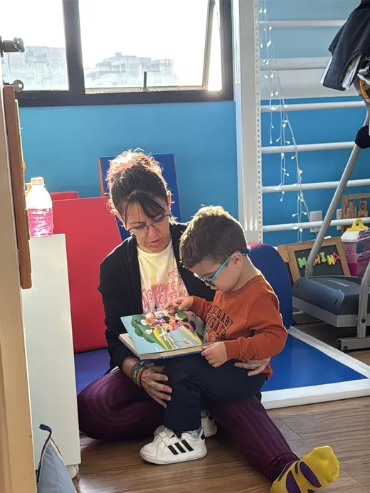 Dafne lendo um livro ilustrado com uma criança sentada ao seu lado