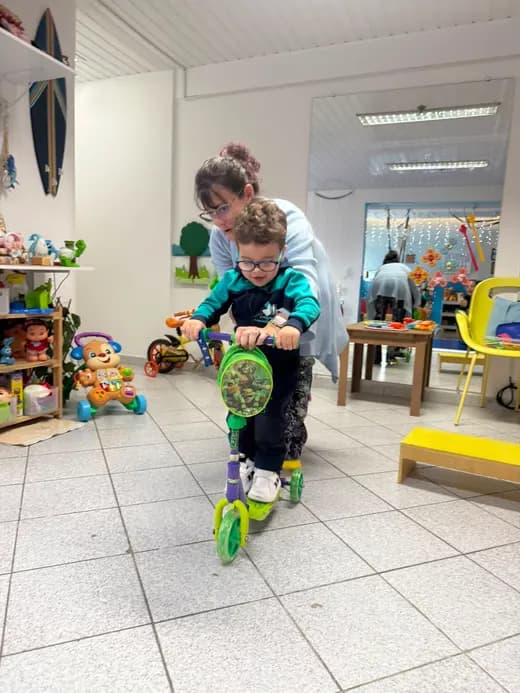 Criança em patinete adaptado numa sala de atendimento, com Dafne atrás