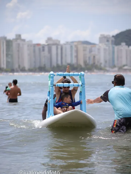Criança em cadeira adaptada entrando no mar, acompanhada pela equipe